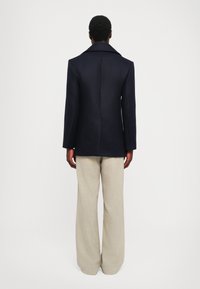 Námornícky kabát z vlnenej zmesi s výstrihom do V, priliehavým strihom a štruktúrovanými plecami, v kombinácii s svetlo béžovými širokými nohavicami.