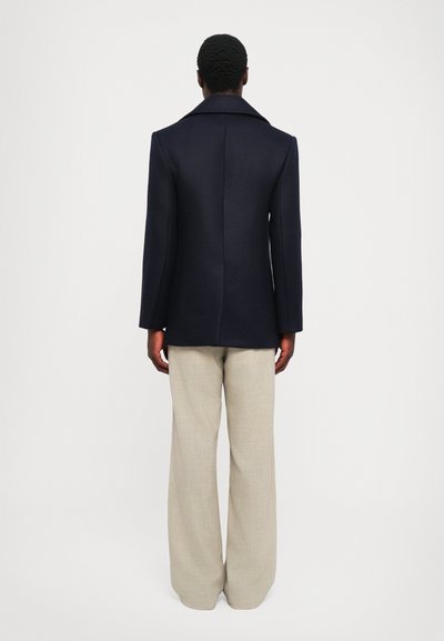 Jūras spēku tumši zils vilnas maisījuma mētelis ar izgrieztu apkakli, pielāgotu dizainu un strukturētām plecu daļām, savienots ar gaiši bežām platām biksēm.