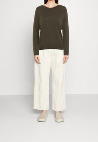 Person, die ein langärmliges, dunkelolivfarbenes Oberteil, lockere weiße Hosen und beige Slipper trägt, steht vor einem schlichten hellen Hintergrund.