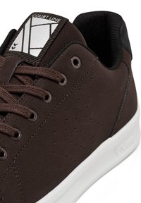 Brązowy sneaker o gładkiej, matowej teksturze. Posiada perforacje po bokach, brązowe sznurowadła i białą gumową podeszwę; zawiera szary język z logo.