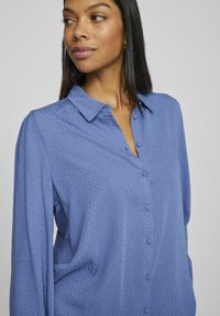 Lichtblauwe blouse met knopen, gemaakt van een zachte, gestructureerde stof, met een kraag, lange mouwen en subtiele stippenpatronen.}}}