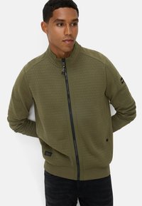 Olivengroene,ritsjack met honingraat textuur, hoge kraag en contrasterende zwarte rits. Bevat een logo patch op de mouw.