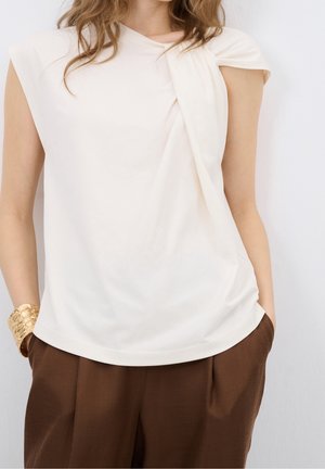 Femme portant un haut blanc sans manches drapé et un pantalon marron, les mains dans les poches et un bracelet manchette doré au poignet gauche.