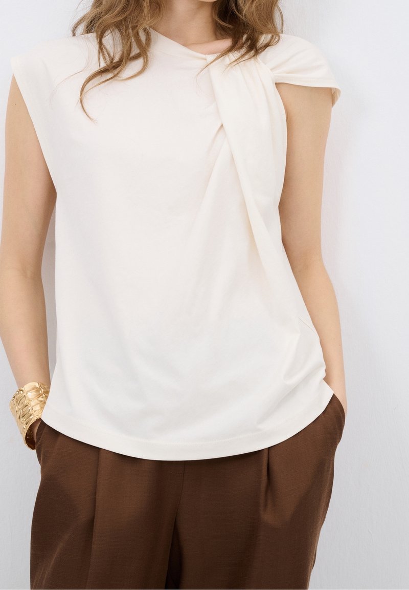 Femme portant un haut blanc sans manches drapé et un pantalon marron, les mains dans les poches et un bracelet manchette doré au poignet gauche.