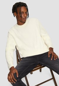 Hellbrauner gerippter Strickpullover mit Rundhalsausschnitt, kombiniert mit dunkelgrauen Jeans. Model sitzt auf einem Holzstuhl.