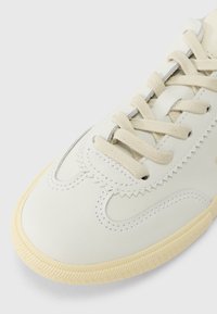 Λευκό αθλητικό παπούτσι από δέρμα με κρεμ κορδόνια, διακοσμητικές ραφές, στρογγυλεμένη μύτη και υφασμένο ελαστικό πέλμα σε ανοιχτό μπεζ χρώμα.