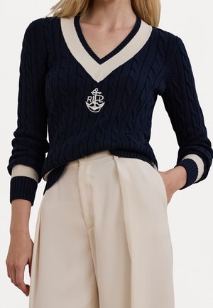 Mujer con un suéter de punto trenzado azul marino con cuello en V, ribete blanco y emblema de ancla, combinado con pantalones beige de pierna ancha y tiro alto.