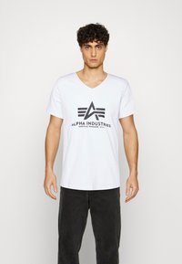 Bílé tričko s výstřihem do V vyrobené z bavlny s černým logem obsahujícím "Alpha Industries" a třemi pruhy nad ním. Nošeno s tmavými kalhotami.