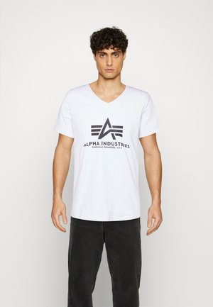 Hvid v-hals t-shirt lavet af bomuld med et sort logo, der indeholder "Alpha Industries" og tre striber ovenfor. Bærer med mørke bukser.