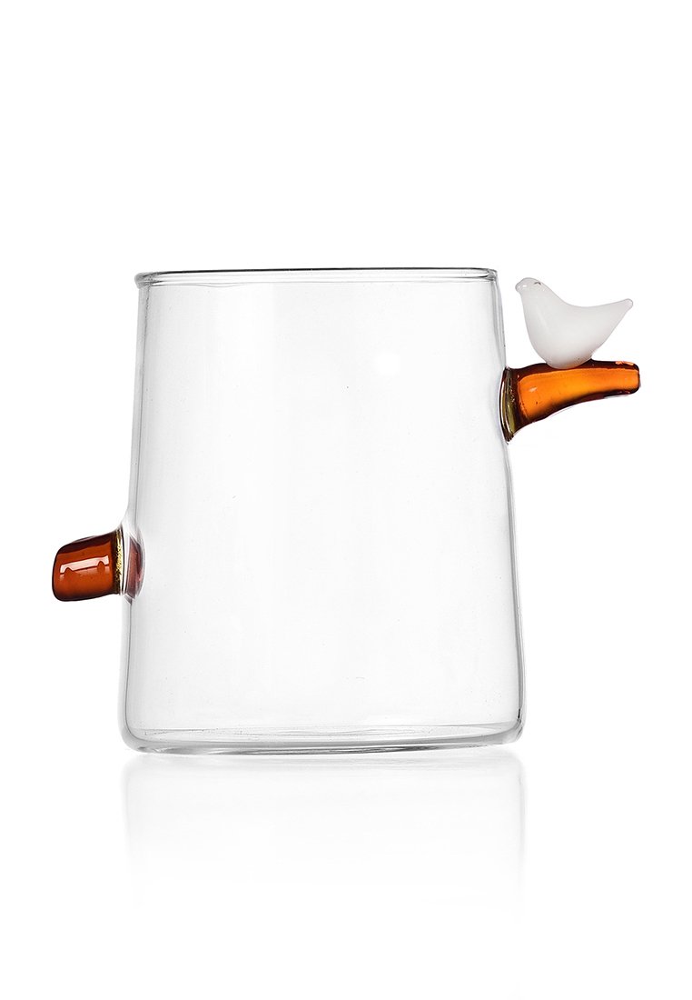 Przezroczysty szklany kubek z dwoma bursztynowymi uchwytami, z których jeden zwieńczony jest małą białą figurką ptaka, na białym tle.