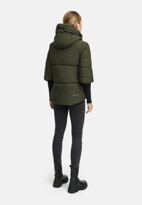 Padded, olive green jacket with a hood, short sleeves, and a textured finish. Features a logo label at the back. Worn over black pants and boots.