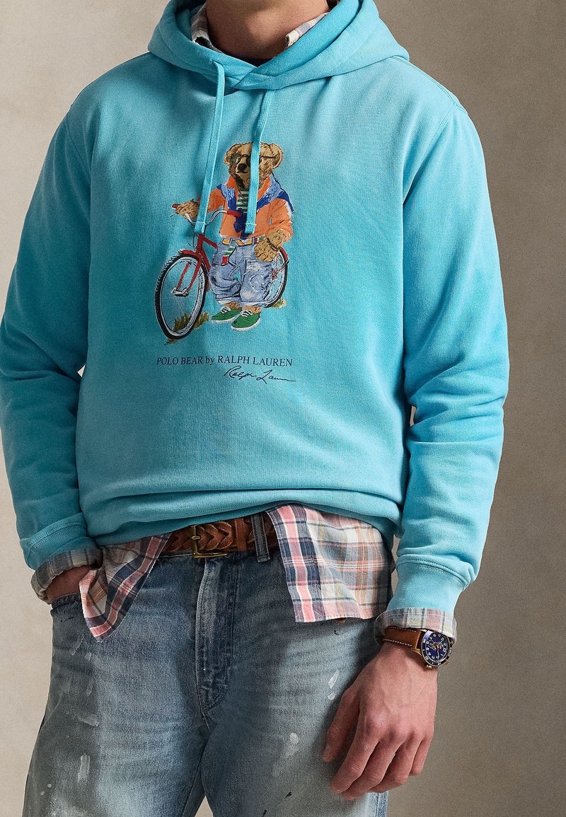 Homme portant un sweat à capuche bleu clair avec un dessin d'ours en train de faire du vélo, superposé à une chemise à carreaux, accompagné d'un jean délavé et d'une montre.