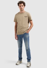 Jonge man met krullend haar staat tegen een effen achtergrond, draagt een beige graphic t-shirt, blauwe spijkerbroek en zilveren sneakers, met zijn hand in zijn zak.