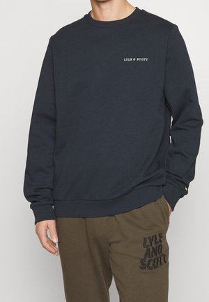 Homme portant un sweat-shirt bleu marine avec le texte « LYLE & SCOTT » sur la poitrine et un pantalon vert olive avec le texte « LYLE AND SCOTT » sur la cuisse.
