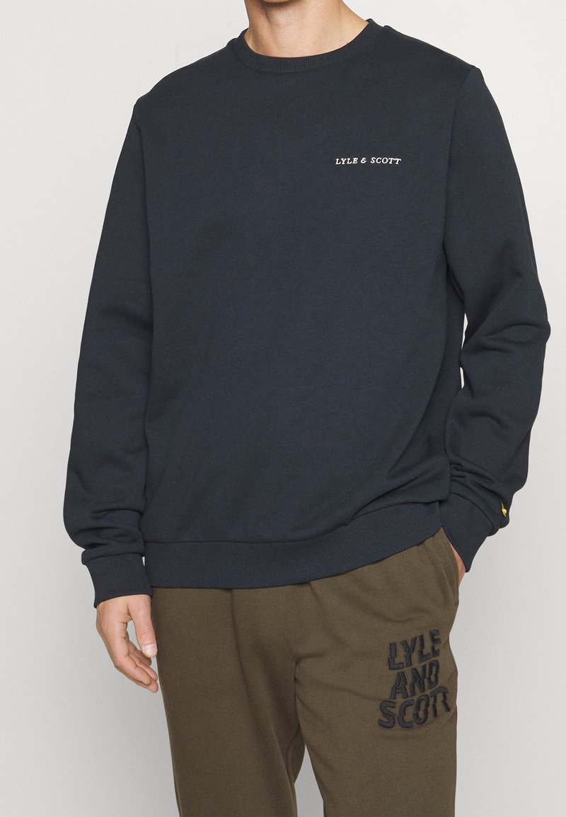Homme portant un sweat-shirt bleu marine avec le texte « LYLE & SCOTT » sur la poitrine et un pantalon vert olive avec le texte « LYLE AND SCOTT » sur la cuisse.