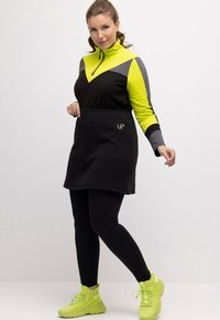 Zwarte sportrok en lange mouwen top met neon gele en grijze accenten. Heeft een ritskraag en logodetail. Draag het met zwarte leggings en heldere sneakers.
