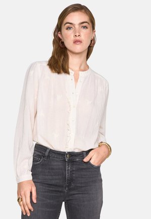 Blusa bianca con scollatura rotonda e dettagli di ruffles sul davanti, abbinata a jeans scuri. La blusa presenta maniche lunghe e leggermente a sbuffo.