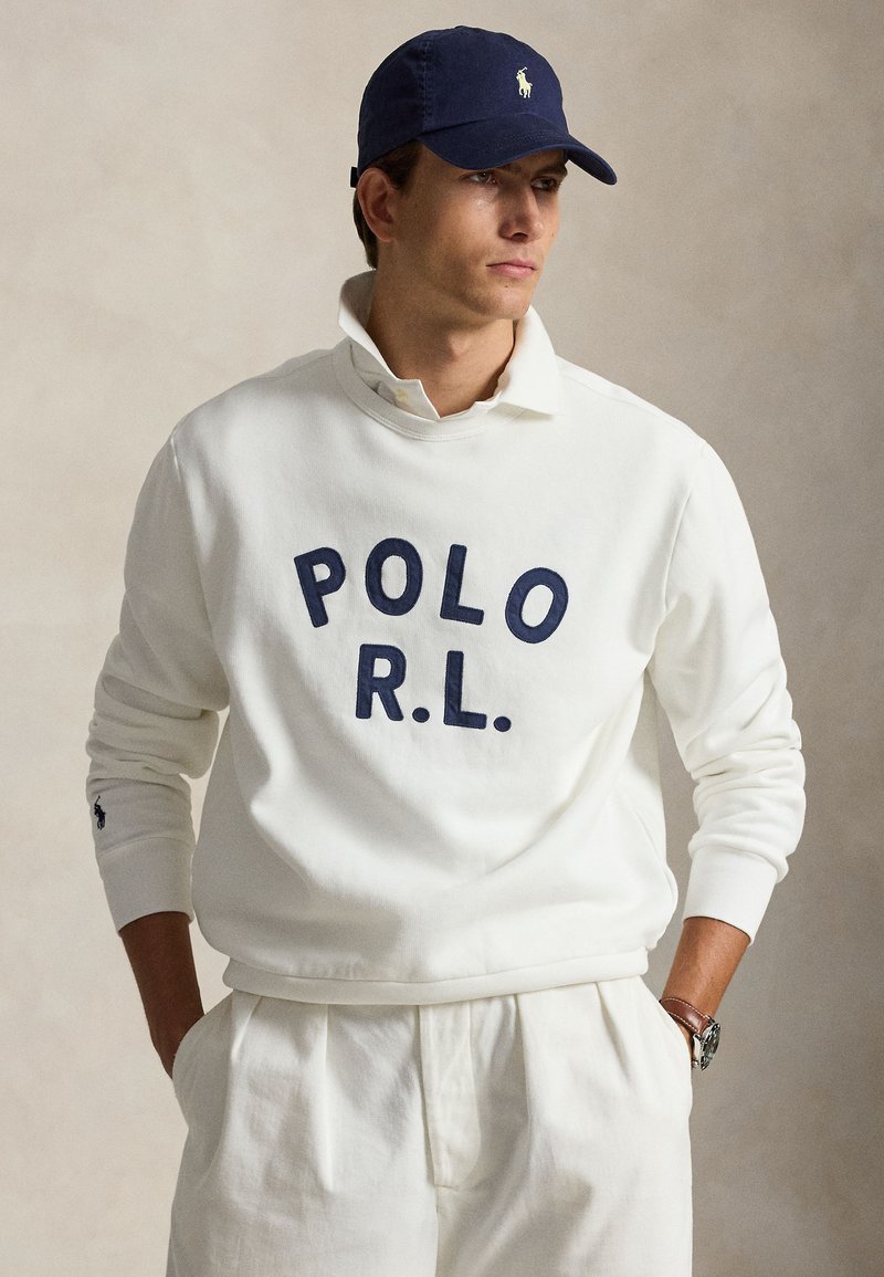 Άνδρας με λευκό φούτερ Polo Ralph Lauren, λευκό παντελόνι, μπλε σκουφάκι με λογότυπο και ρολόι, στέκεται μπροστά σε απλό φόντο.