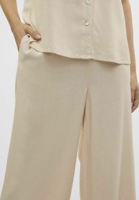 Beige vida byxor med en mjuk textur och subtil lyster. Har sidofickor och en avslappnad passform, kombinerat med en matchande knappad topp.
