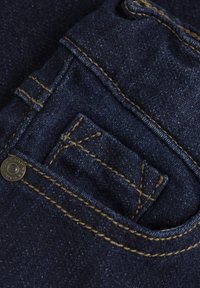 Mörkblå denimtyg med en liten myntficka med gul sömnad och en metallknapp, som framhäver texturen och slitaget på materialet.