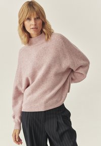 Jasnoróżowy sweter dziany z wysokim dekoltem, opadającymi ramionami i ściągaczami, połączony z ciemnymi spodniami w prążki.