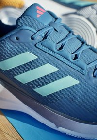 Ingrandimento di una scarpa sportiva blu con strisce azzurro chiaro, lacci azzurro chiaro e un logo rosso su una superficie di legno.