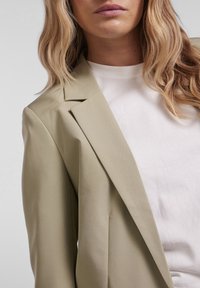 Femme aux cheveux blonds ondulés portant un blazer beige sur un haut blanc à col rond, sur un fond gris clair.