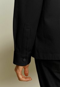 Zwarte textuurjas met een gerafelde manchetknoop, met een opvallend naaddetail en een rechte snede aan de mouw. Hand gedeeltelijk zichtbaar.