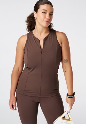 Femme tenant une raquette de paddle blanche et dorée, portant un haut marron sans manches et des leggings, avec des cheveux bouclés attachés en arrière et de petits tatouages sur le bras.