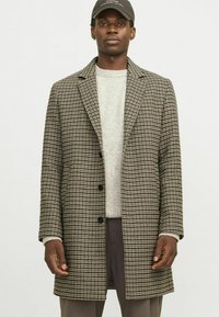 Pruun ja must houndstooth mustriga mantel, millel on nurgelised kraed, kolm nuppu ja küljetaskud, kantud heledat halli sviitrit kandes.