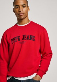 Sudadera roja hecha de una mezcla de algodón con cuello redondo, que presenta el texto negro "PEPE JEANS" y "Londres 1973" en una fuente negrita y curva.