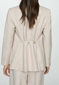 Blazer rose pâle sur mesure avec des rayures verticales, cintré à la taille et avec des poignets boutonnés. Tissu lisse avec une coupe structurée et formelle.