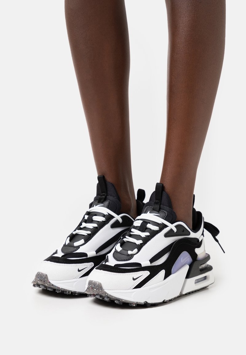 Jambes portant des baskets Nike noires et blanches avec des semelles épaisses, des lacets blancs et un détail violet sur les côtés, debout sur une surface blanche.