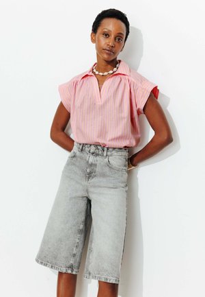 Femme debout sur fond blanc portant une chemise à manches courtes rayée rose, un short en jean gris jusqu'aux genoux, un collier en coquillages et un bracelet.