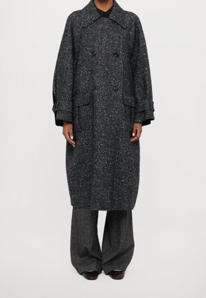 Persoon die een lange, donkergrijze gemêleerde wollen jas draagt met brede kraag, grote knopen en bijpassende wijde grijze broek.