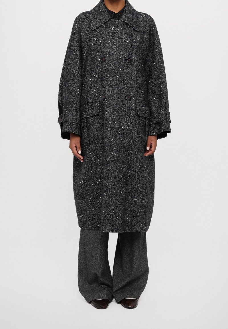 Personne portant un long manteau en laine gris foncé moucheté avec un large col, de gros boutons, et un pantalon gris à jambes larges assorti.