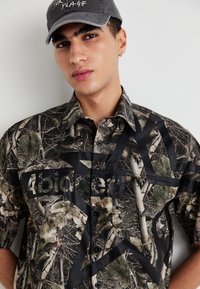Cămașă scurtă, cu model camuflat în nuanțe de verde și maro, având un print grafic negru și închidere cu nasturi. Guler ajustabil.