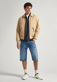 Chaqueta bomber beige con dos bolsillos laterales, superpuesta sobre una camiseta gris oscuro. Combinada con pantalones cortos de mezclilla azul claro y zapatillas blancas con detalles en marino.