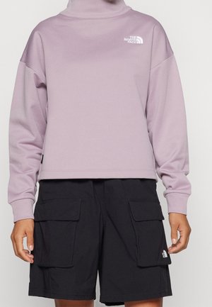 Personne portant un sweat-shirt à col montant lilas à manches longues et un short cargo noir avec poches avant à rabat, les deux avec les logos The North Face.