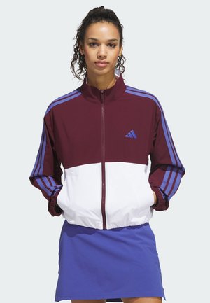 Femme portant une veste zippée bordeaux et blanche avec des rayures bleues et une jupe bleue, debout les mains dans les poches de la veste.