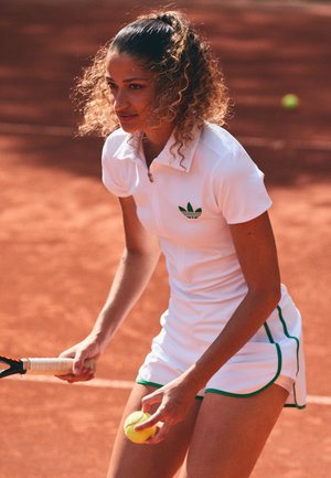 Vestido atlético blanco con cuello, acentos verdes y logo; el jugador sostiene una pelota de tenis, preparado en una cancha de tierra batida.