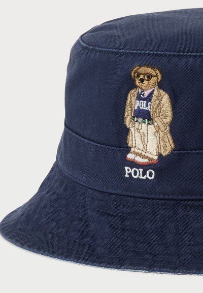 Chapeau seau bleu marine en coton avec une finition texturée. Présente un ours brodé en costume avec le texte "POLO" en dessous.