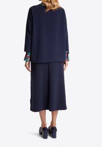 Frau in navy-blauen, weit geschnittenen, verkürzten Hosen, passender lockerer Jacke, floralem Hemd mit aufgeschlagenen Ärmeln und navy-blauen Blockabsatzschuhen, stehend mit dem Rücken zur Kamera.