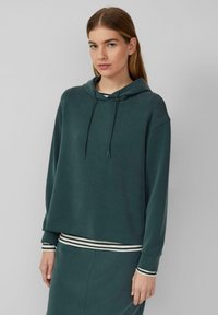 Jauna sieviete, ģērbusies teal krāsas kapucē un saskaņoto svārku, ar svītrainām krekla piedurknēm, stāvot pret vienkāršu gaiši pelēku fonu.