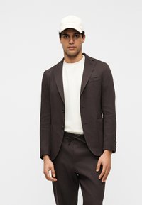 Άνδρας που φοράει σκούρο καφέ σακάκι και παντελόνι με λευκό μπλουζάκι και λευκό καφέ μπέιζμπολ καπέλο, μπροστά σε απλό φόντο.