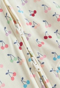 Gros plan sur un tissu crème avec des motifs de cerises multicolores et deux tirettes de fermeture éclair en plastique blanc partiellement fermées.