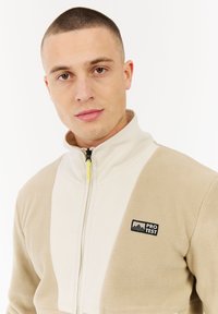 Beige en crème fleec jasje met een ritssluiting aan de voorkant, een hoge kraag en een zwart logo op de linkerborst. Gladde textuur en atletische pasvorm.
