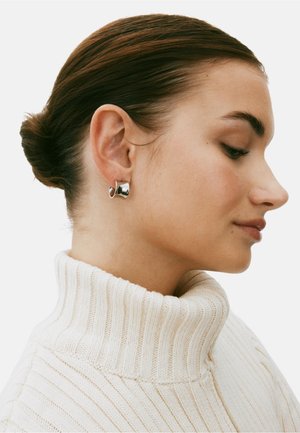 Silberne Creolen mit einem geschwungenen Design, präsentiert an einem Modell mit einem hellbeigen gerippten Rollkragenpullover. Glatte metallische Textur.