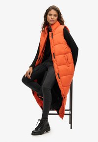 Lange, puffer vest in fel oranje met een hoge kraag, voorzakken en zwarte accenten, gedragen over een zwart topje en jeans, gecombineerd met zwarte laarzen.