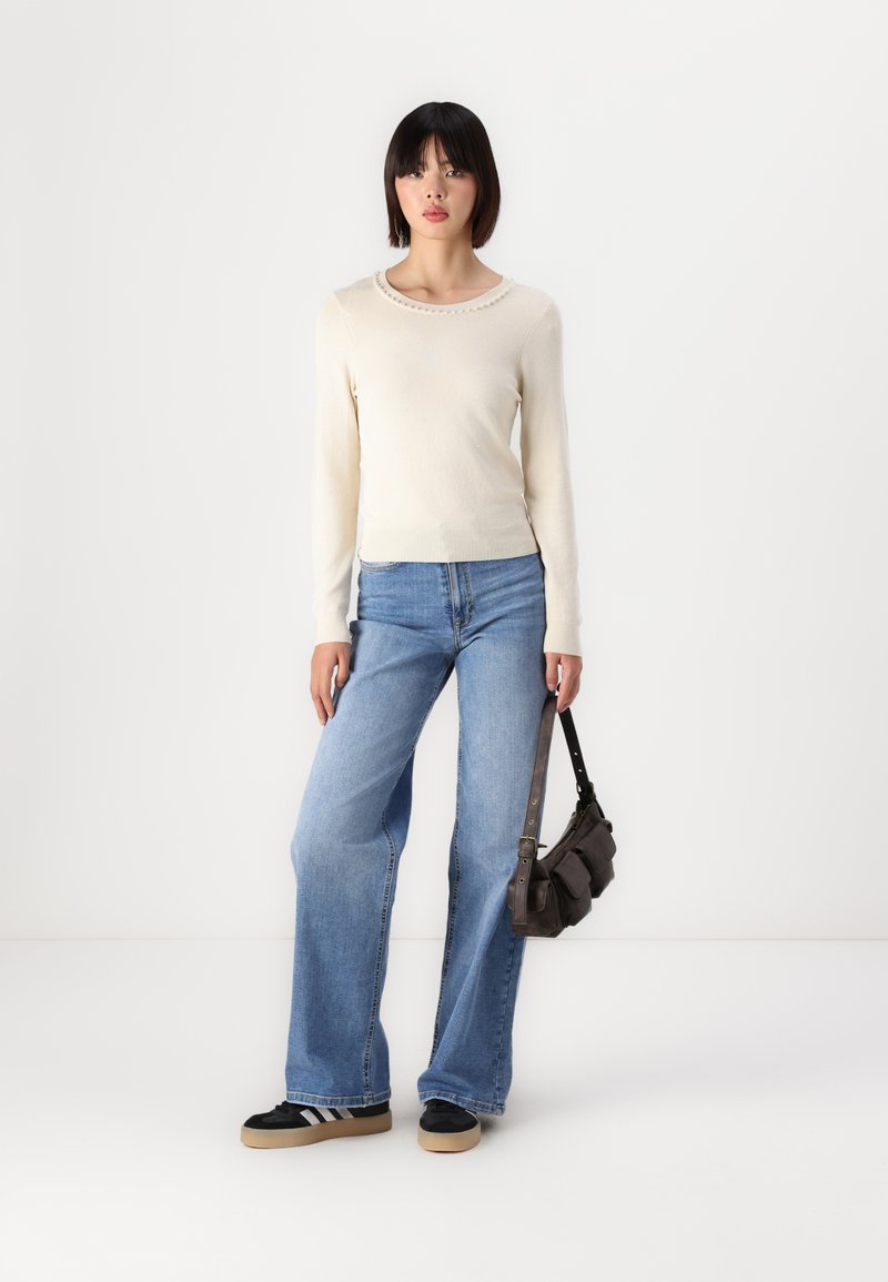 Maglione a maniche lunghe in maglia crema con scollatura ondulata, abbinato a jeans in denim a gamba larga di colore blu chiaro e sneakers nere con strisce bianche.